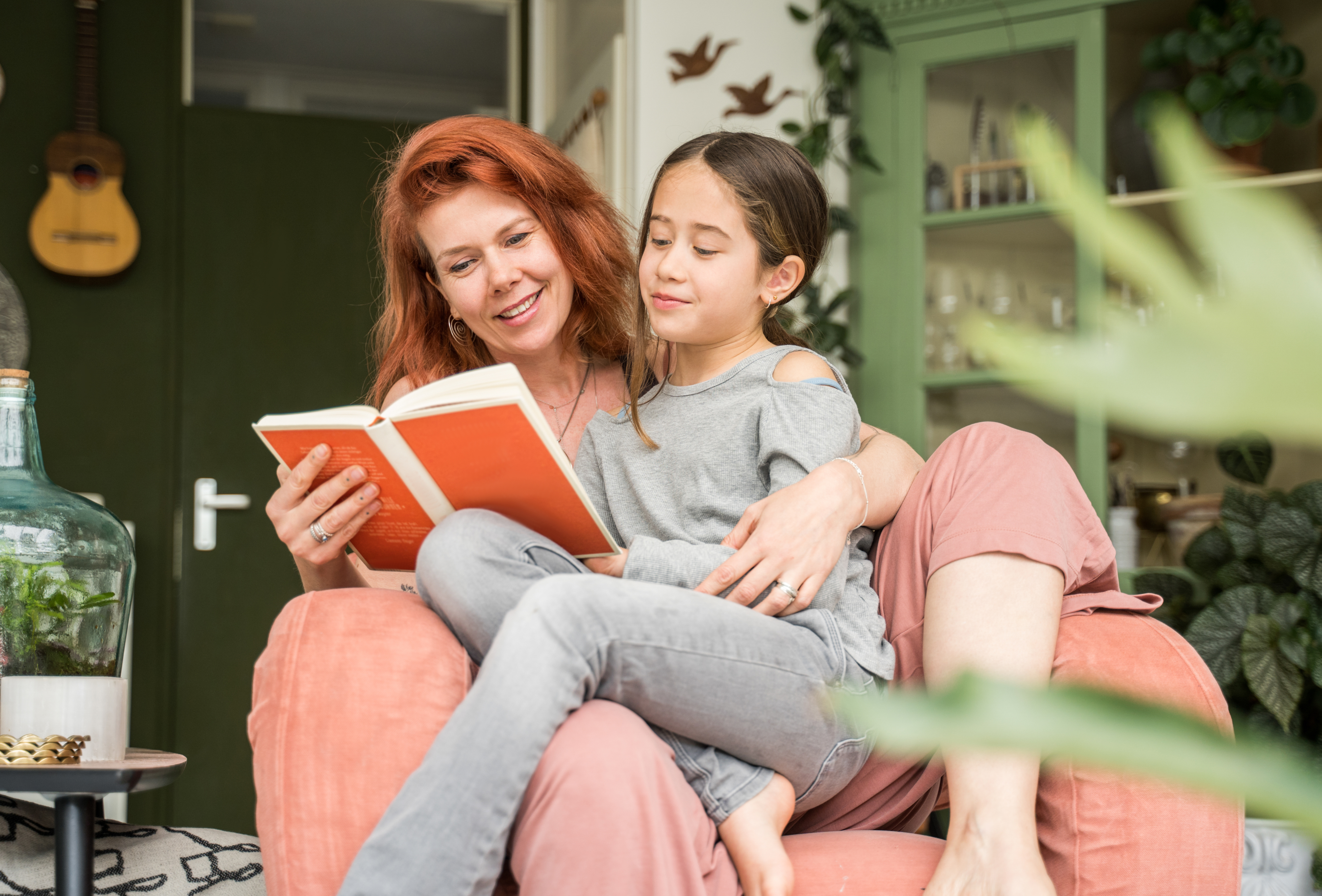 Eine Mutter liest ihrer Tochter im Grundschulalter aus einem Buch vor. Das Mädchen sitzt auf ihrem Schoss. 