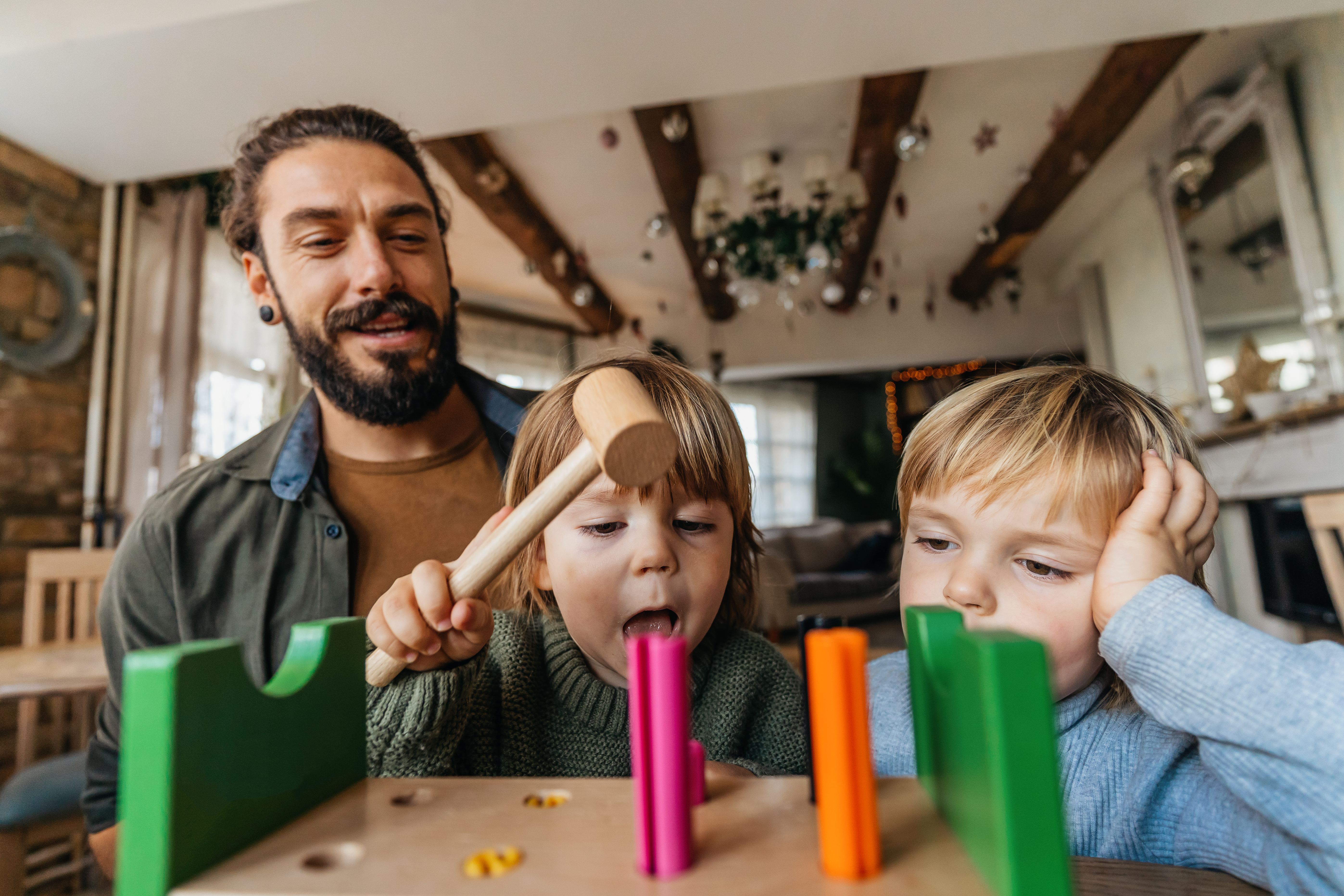 Ein bärtiger Mann spielt gemeinsam mit zwei Kindern im Kleinkindalter. Sie spielen mit einem Holzbaukasten. 